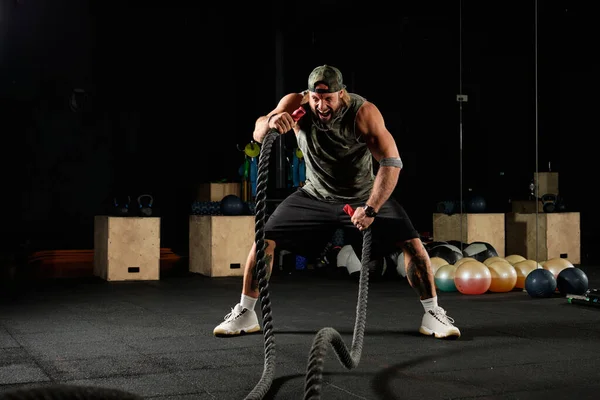 Hombre musculoso con un uniforme deportivo y una gorra de béisbol en un estante realizando violentamente un ejercicio con cuerdas — Foto de Stock