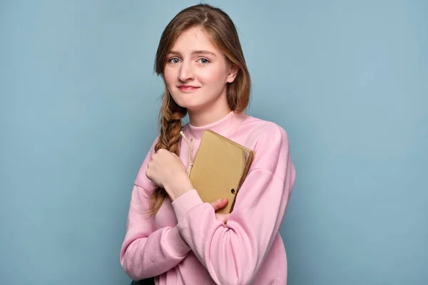 En tjej i rosa tröja kramar en bok och väskor sina läppar medan hon tittar på kameran. — Stockfoto