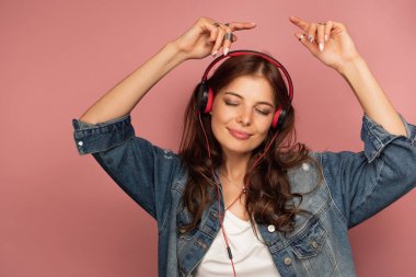 Pembe arka planda esmer, kot pantolon ve kulaklıkla dans ediyor gözlerini kapatıyor ve kollarını başının üstüne kaldırıyor..