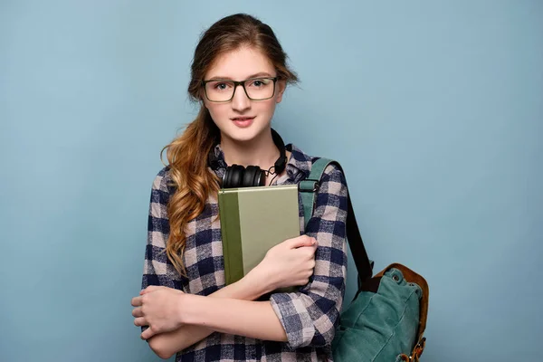 En flicka i rutig skjorta och glasögon med hörlurar på halsen står med en ryggsäck, håller en bok för sig själv. — Stockfoto