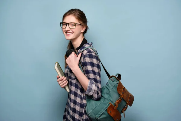 Skrattande flicka i rutig skjorta och glasögon på en blå bakgrund med en bok och en ryggsäck. — Stockfoto