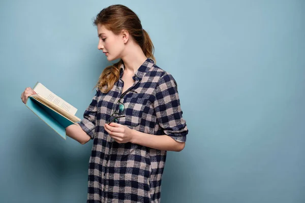 En blond flicka i rutig skjorta står i profil på en blå bakgrund och läser en bok med glasögon i handen. — Stockfoto