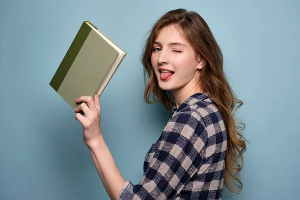 En ung flicka i rutig klänning står på en blå bakgrund blinkar och sticker ut tungan, håller en bok i handen. — Stockfoto