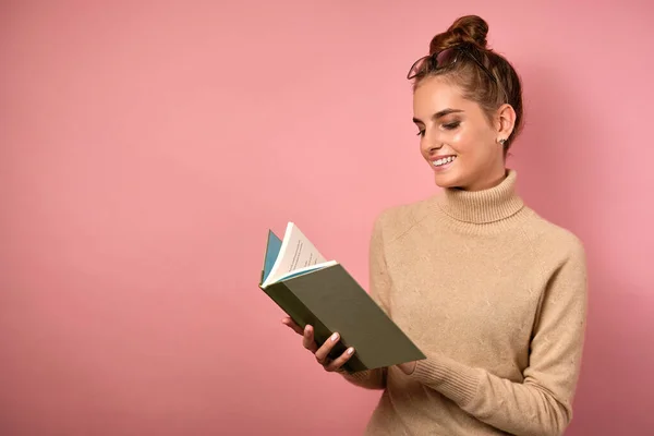 머리와 안경을 이마에 쓴 채 핑크 색 배경 위에 서 있는 베이지 색 터틀넥을 입은 한 소녀가 열린 책을 보고 있습니다. — 스톡 사진