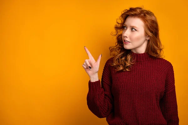 Rödhårig lockig flicka i en vinröd tröja står på en gul bakgrund, blickar åt sidan, pekar med fingret. — Stockfoto