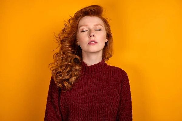 노란 배경에 앉아 눈을 감은 채 헐렁 한 스웨터를 입고 있는 빨간 머리 소녀가. — 스톡 사진