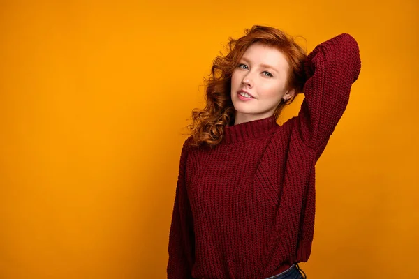 Egy vörös hajú, göndör lány egy burgundi pulóverben sárga alapon áll, és belenéz a keretbe a kezével a feje mögött. — Stock Fotó