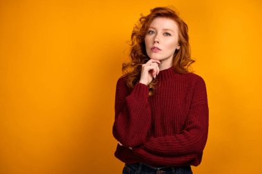 Red-haired girl in a burgundy sweater is standing on an orange background and propping chin with her hand, looks into the frame