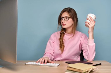 Pembe kazaklı ve gözlüklü bir kız bilgisayarın önünde oturmuş elinde kağıt bardakla monitöre bakıyor..