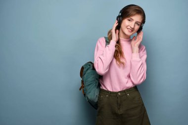 Pembe kazaklı, sırt çantalı bir kız mavi arka planda duruyor ve kulaklık tutuyor..
