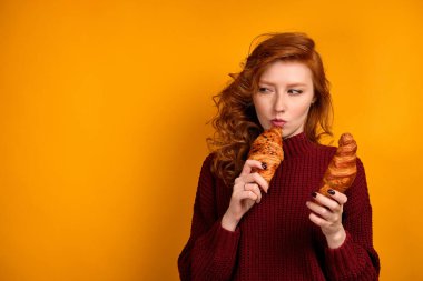 Kırmızı saçlı, bordo kazaklı bir kız turuncu arka planda duruyor ve dudaklarına kruvasan koyuyor.