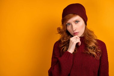 Burgonya rengi süveter giyen, turuncu arka planda duran kıvırcık kızıl saçlı bir kız çenesine tutunarak çerçeveye bakar..