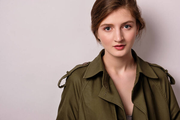 Девушка в зеленом пальто с собранными волосами стоит на светлом фоне и смотрит в раму
