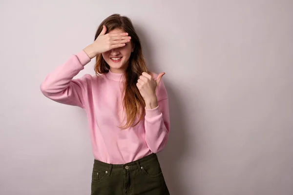Flickan i rosa t-shirt står på en ljus bakgrund, ler och blundar med en hand pekar tummen åt sidan. — Stockfoto