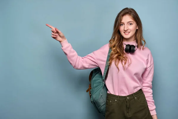 Chica en una sudadera rosa con una mochila y auriculares sobre un fondo azul, señala un dedo a un lado — Foto de Stock