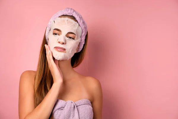 Dziewczyna w liliowym ręczniku stoi z maską na twarzy na różowym tle. — Zdjęcie stockowe