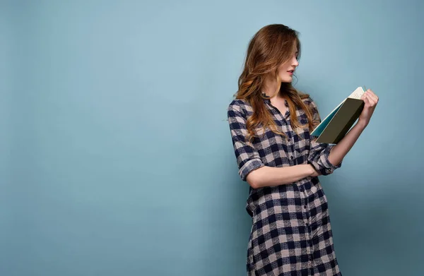 En flicka i rutig skjorta står i en halvsväng på en blå bakgrund och läser en bok. — Stockfoto