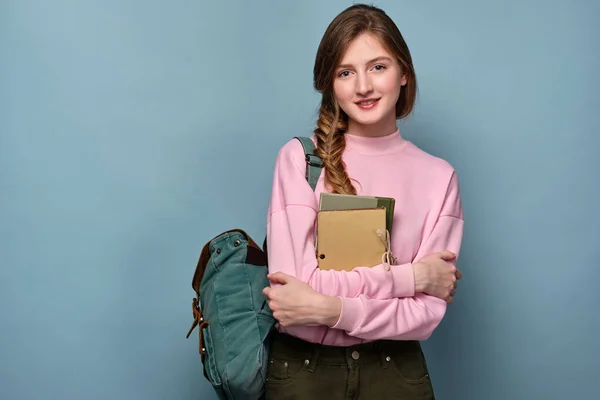 Egy kék szemű lány rózsaszín pulóverben kék háttéren áll hátizsákkal és könyvekkel a kezében.. — Stock Fotó