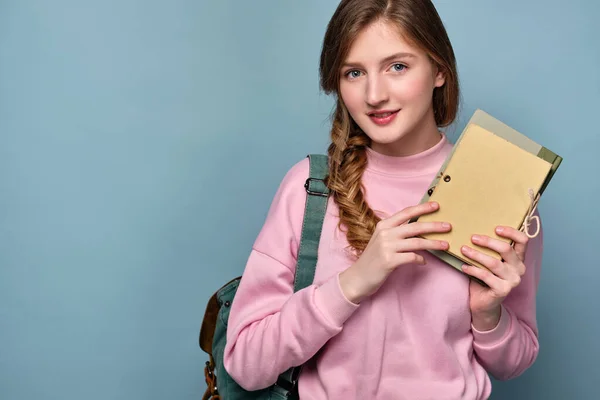 Egy lány rózsaszín pulóverben, copfban, kék háttérben áll egy hátizsákkal, jegyzetfüzetekkel és könyvekkel a kezében.. — Stock Fotó