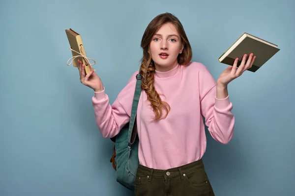 En flicka i rosa tröja med en fläta står med en ryggsäck och distraherat kastar upp händerna med böcker åt sidorna. — Stockfoto