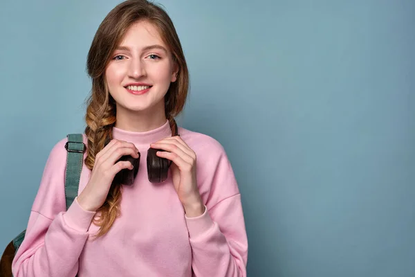 Una chica con ojos azules y una trenza en un suéter rosa sostiene los auriculares en su cuello y sonríe . — Foto de Stock
