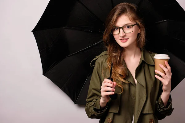 En flicka står mot bakgrunden av ett öppnat svart paraply i en grön trenchcoat, glasögon med pappersmugg i handen — Stockfoto