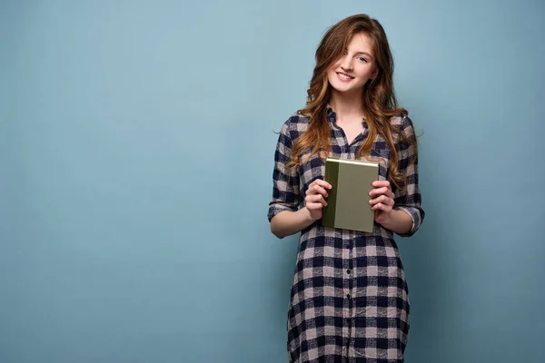 En flicka i rutig skjorta står med en bok i händerna på en blå bakgrund och ler in i ramen — Stockfoto