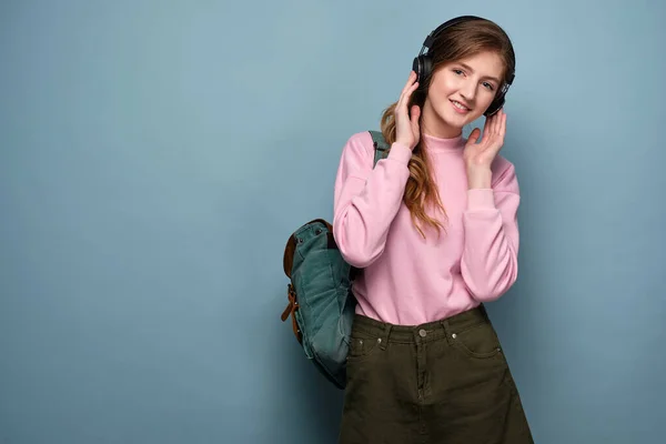 En flicka i rosa tröja med en ryggsäck står på en blå bakgrund och håller hörlurar. — Stockfoto