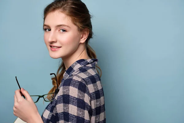 En tjej med blå ögon i rutig skjorta är halvvriden och tittar på kameran med glasögon i händerna. — Stockfoto