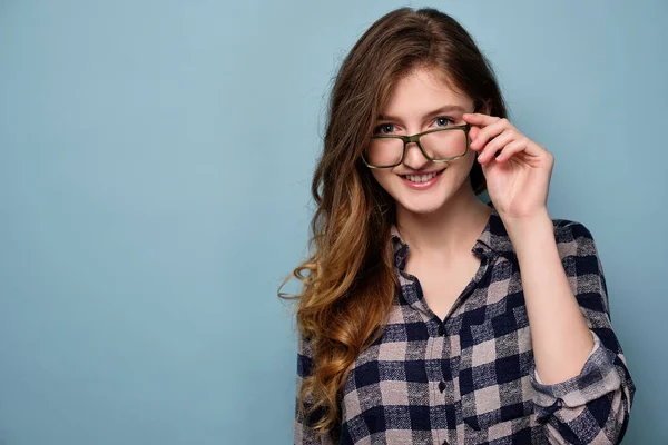 Uma menina em uma camisa xadrez fica em um fundo azul e sorri, segurando seus óculos para baixo . — Fotografia de Stock
