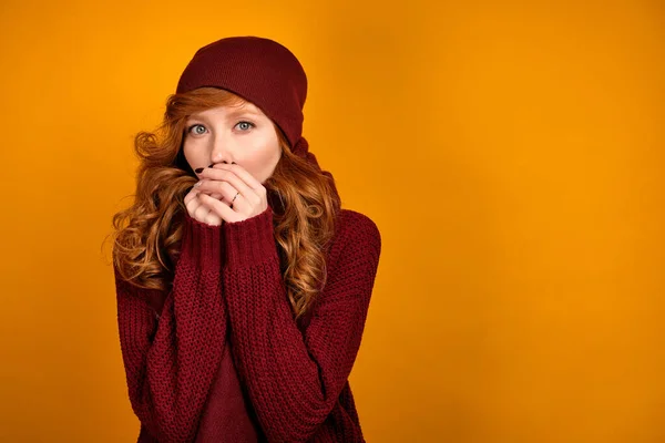 Egy göndör vörös hajú lány burgundi pulcsiban, sállal és sapkával narancssárga alapon áll, és felmelegíti a kezét, belélegzi őket. — Stock Fotó