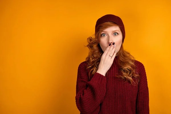 En rödhårig flicka i en vinröd tröja och hatt står på en orange bakgrund, förvånansvärt öppna ögon, täcker munnen med palm — Stockfoto