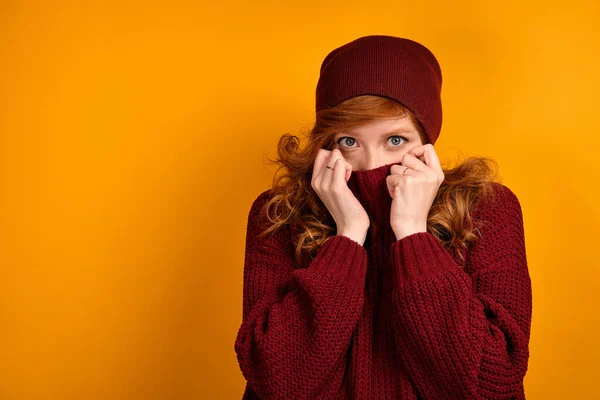 Lockigt rödhårig flicka i en vinröd tröja och hatt, står på en orange bakgrund, lyfta kragen till ögonen. — Stockfoto