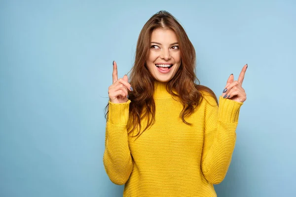 Eine schöne Brünette im gelben Pullover steht auf blauem Hintergrund, zeigt mit den Fingern nach oben und schaut zur Seite — Stockfoto