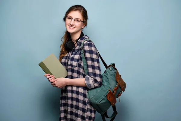 Flicka i rutig skjorta och glasögon står halvvänd på en blå bakgrund med en ryggsäck och en bok i händerna, ler. — Stockfoto