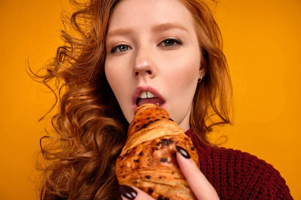 Una chica de pelo rojo rizado en un suéter de color burdeos está de pie sobre un fondo naranja y muerde un croissant, vista desde abajo —  Fotos de Stock