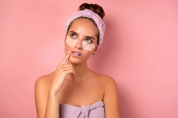 A girl in a towel with a bandage on head and patches under eyes, looks thoughtfully at the top, propping chin with finger — Stock Photo, Image