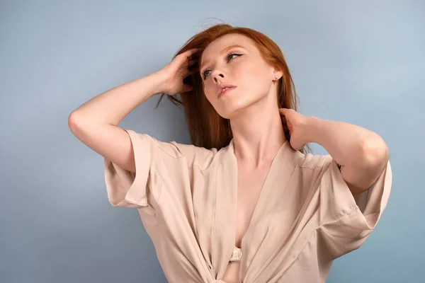 Una chica pelirroja con una túnica ligera se levanta sobre un fondo azul y levanta la barbilla, mira a un lado, alisándose el pelo . —  Fotos de Stock