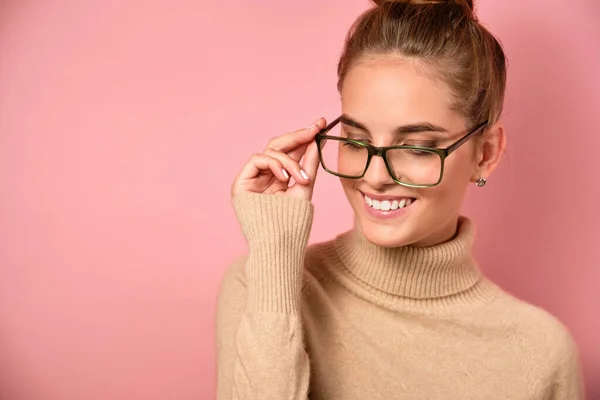 Una chica con una viga alta está de pie en un suéter sobre un fondo rosa, pone gafas en el puente de la nariz, se centran en las gafas — Foto de Stock