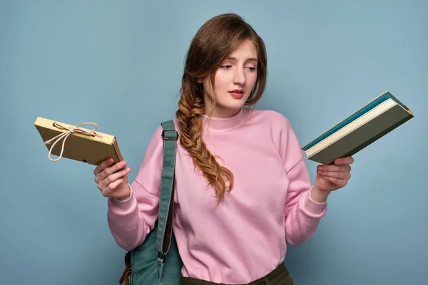 En flicka i rosa tröja med en fläta står på en blå bakgrund med en ryggsäck och tittar på förvirrade böcker i händerna. — Stockfoto