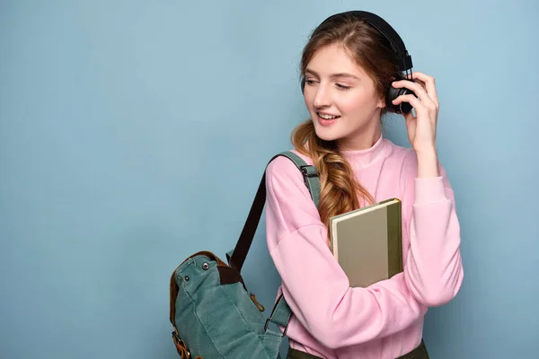 En flicka i rosa tröja står på en blå bakgrund med en ryggsäck och bok, justera sina hörlurar och titta åt sidan — Stockfoto