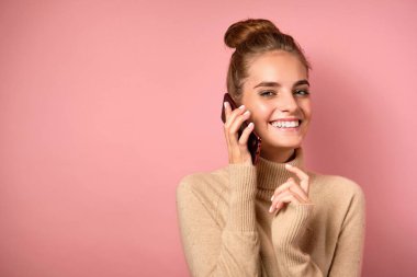 Yüksek kirişli bir kız pembe arka planda süveter giyer, akıllı telefonu kulağından tutar ve çerçeveye gülümser..