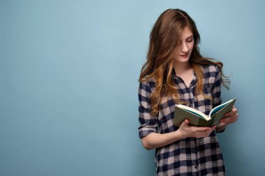 Ekose gömlekli sarı saçlı bir kız mavi arka planda duruyor ve bir kitap okuyor..