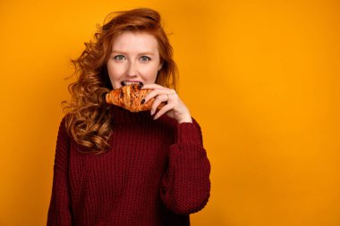 Burgonya rengi kazaklı kıvırcık saçlı bir kız turuncu bir arka planda duruyor ve çerçeveye bakıyor ve bir kruvasanı ısırıyor..