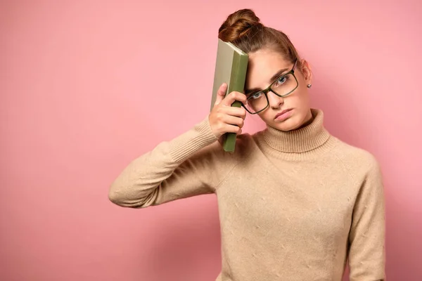 En flicka i en polotröja och glasögon med samlat hår står på en rosa bakgrund med en torterad ansikte, hålla en bok för att huvudet — Stockfoto