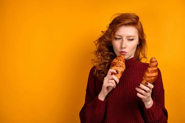 Egy göndör vörös hajú lány burgundi pulóverben croissant-t tart, és figyelmesen néz az egyikre.. — Stock Fotó