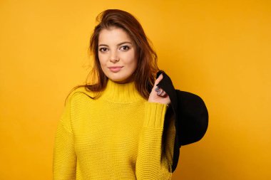 Sarı kazaklı güzel bir esmer sarı arka planda duruyor ve siyah bir şapka tutuyor, çerçeveye bakıyor..