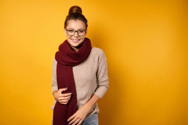 Gözlüklü bir esmer ve yüksek kirişli bordo bir eşarp, sarı bir arka planda durur ve çerçeveye gülümser..