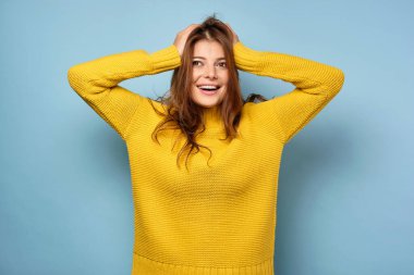 The brunette stands on a blue background in a yellow sweater and looks to the side, ruffling her hair with her hands. clipart