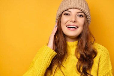 Sarı süveterli ve şapkalı bir esmer sarı arka planda duruyor ve saçlarını düzeltirken çerçeveye gülümsüyor..